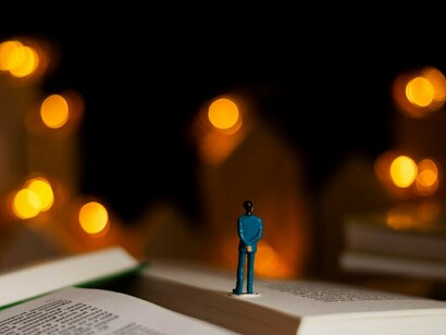 A still life of books and imagination, featuring a human figure atop the stacked volumes
