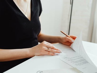 Uma mulher segura e organiza documentos sobre uma mesa, ocupando sozinha o espaço diante dos papéis que precisam ser preenchidos e conferidos. A imagem dialoga com a trajetória narrada pelo autor, marcada pela presença materna diante das responsabilidades e pelo silêncio em torno de uma figura paterna ausente