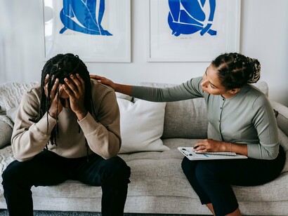 Uma mulher acolhe um homem visivelmente abatido em um momento de vulnerabilidade sentados em um sofá. O apoio emocional e a escuta podem ajudar a reconhecer sinais que o corpo expressa quando as emoções permanecem silenciadas