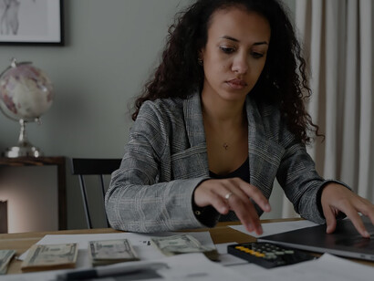Amidst the blend of tradition and technology, an African woman carefully ponders financial decisions, seated before a tableau of coins and a computer in her office