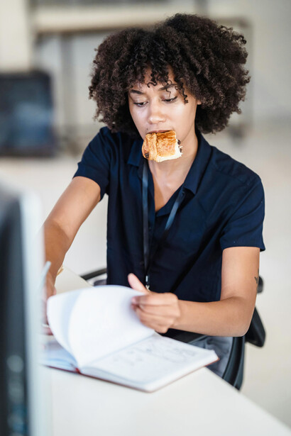 A portrait of a woman multitasking with paperwork while eating a croissant in an office setting, reflecting stress and anxiety