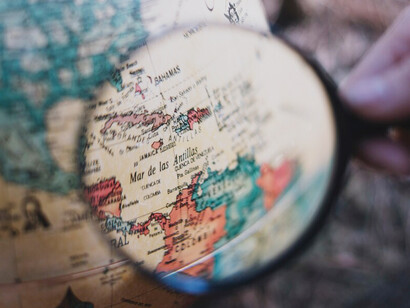 A person using a magnifying glass to closely inspect a globe, representing an in-depth investigation of global dynamics and cultural connections