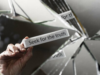 A broken mirror splintering reflections while a hand grips a sheet marked “Seek the truth,” symbolising the fragmentation of truth under profit-driven narratives