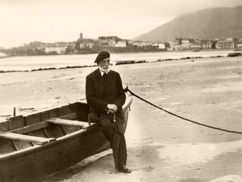 Miguel de Unamuno sentado en barca con boina y paraguas. Mar y pueblo al fondo