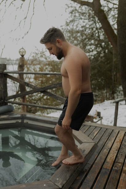 A man carefully steps into a wooden hot tub, contrasting the warmth of the water with the crisp winter air, an example of heat therapy that promotes relaxation, improves cardiovascular health, and supports detoxification
