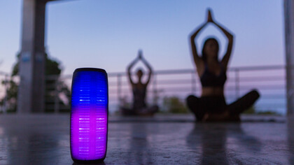 A colorful backlit wireless speaker in the foreground, as a group of friends practices yoga in the background