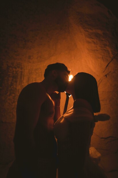 Secret lovers share a tender kiss by torchlight, hidden away in the shadows of a cave