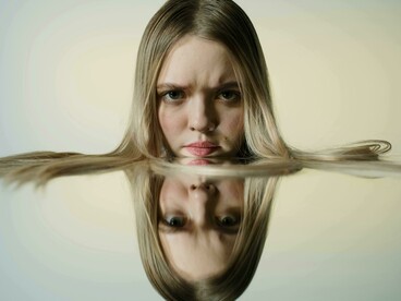 A blonde-haired woman rests her face on a mirror, her uneasy expression symbolising emotional conflict and hypocrisy