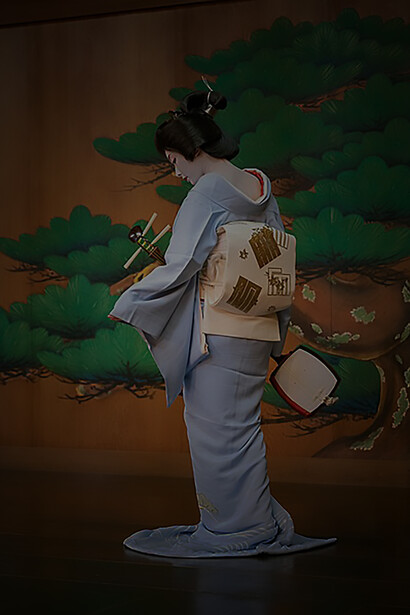 Kyoto geiko Toshimana graces Kawamura Noh Stage, adorned in full make-up and a katsura wig, while elegantly holding a shamisen. Her attire features a stunning blue hikizuri kimono with short sleeves, accentuated by a stiff brocade obi
