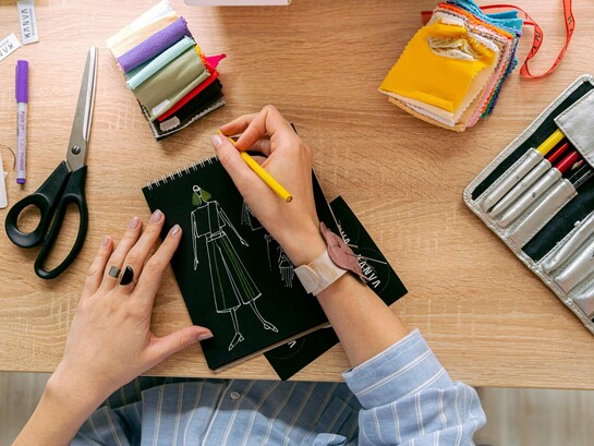 Mãos desenhando roupa em um caderno sobre uma mesa de estudos de moda. Entre traços e ideias, nasce a moda em um processo criativo que une arte, técnica e consciência em cada detalhe. O primeiro esboço de uma peça carrega mais que estética, revela pensamento, cultura e intenção. Antes das passarelas, a moda começa no papel, onde criatividade e propósito se encontram
