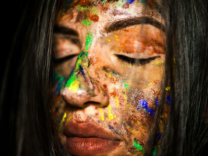A close-up of a woman's face, eyes closed, splattered with colourful paint