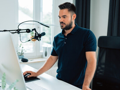 A man in a black t-shirt works diligently at a table in a bustling social media agency, his desk hosts two screens, a central podcast microphone, and a computer displaying a splitscreen interface