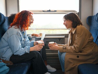 Due amiche si scambiano sorrisi e bevono caffè durante un viaggio in treno, mentre una terza persona legge sullo sfondo