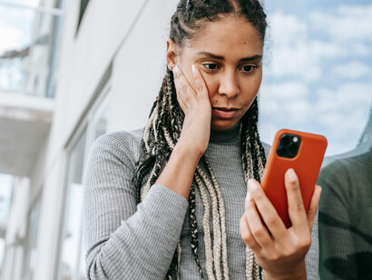 A tired Black woman browses her mobile phone, her expression marked by concern as she stands on the street