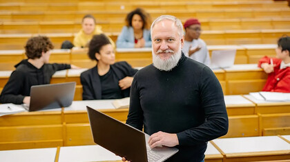A professor wearing a black turtleneck sweater holds a silver MacBook while teaching in a classroom, encouraging active learning at the university