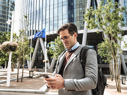 A smiling and successful businessman stands in front of an EU building, using a digital tablet for professional purposes