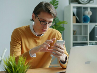Um homem usa o celular enquanto permanece sentado à mesa, cercado por objetos de trabalho. A tela concentra sua atenção e traduz a presença constante da tecnologia no cotidiano, um dos fatores que o artigo associa ao distanciamento da experiência emocional. Em meio à notificações, tarefas e estímulos permanentes, a hiperconectividade pode aproximar o indivíduo do mundo digital e, paradoxalmente, afastá-lo da própria experiência