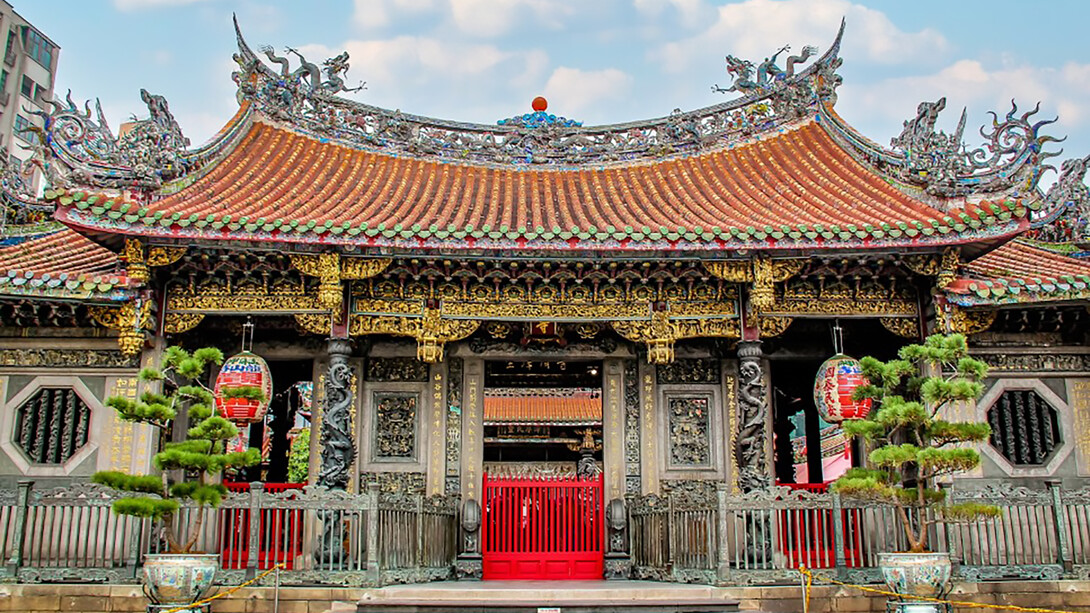 The Longshan Temple in Taipei, Taiwan, features an ancient roof adorned with dragons, set against a backdrop of a clear blue sky and fluffy white clouds