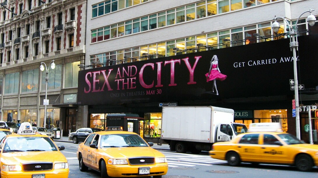 Manhattan em cenário típico de Sex and the City e cartaz em referência à personagem principal.  O seriado contando a história de quatro mulheres solteiras em Nova York atraiu uma legião de fãs, ganhou prêmios e se tornou um marco ao ser um seriado voltado para as mulheres, que falava de sexo e não tinha medo de discutir assuntos considerados tabus na época
