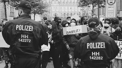 Protesters holding signs that read 'Police Brutality' stand defiantly in front of a line of police officers
