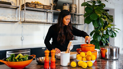 A woman stands gracefully in a kitchen, surrounded by an array of vibrant fruits, while elegantly holding the lid of a pot