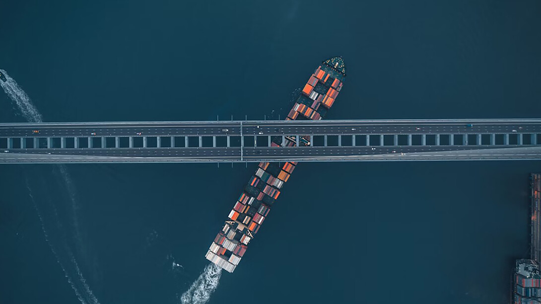 An aerial view of a container cargo ship navigating global trade routes beneath a bridge, symbolizing economic diplomacy and international transportation