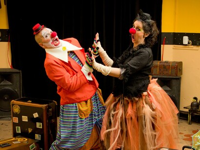 Dois palhaços se apresentam em meio a malas e adereços, em uma cena marcada pelo movimento, pelas cores e pela teatralidade. A brincadeira corporal transforma objetos simples e situações aparentemente banais em possibilidades de jogo, lembrando que, na palhaçaria, o imprevisto e o erro podem deixar de ser obstáculos para se tornar parte essencial da criação