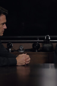A father and son sit across from each other in silence, their tension shaped by control, performance, and the unseen weight of being watched