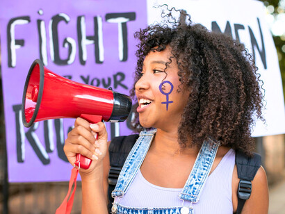A young woman with a feminist symbol painted on her cheek amid protest signs, representing the spirit of resistance and empowerment