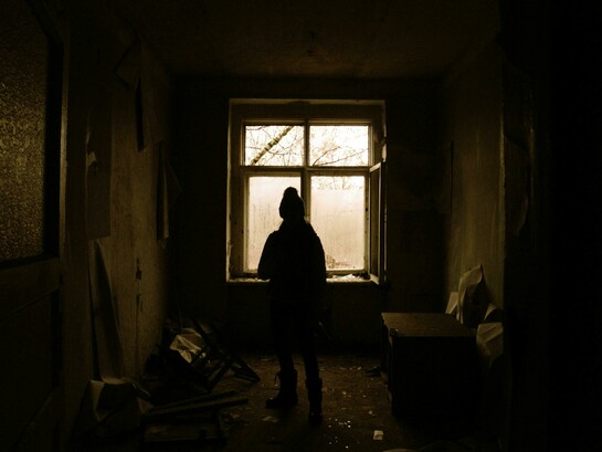 A solitary figure stands silhouetted by a window in an abandoned house, surrounded by fading light and quiet decay
