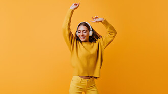 A carefree brunette woman in stylish yellow pants joyfully dancing to a good song, against a backdrop of an inviting orange wall, exuding warmth and happiness