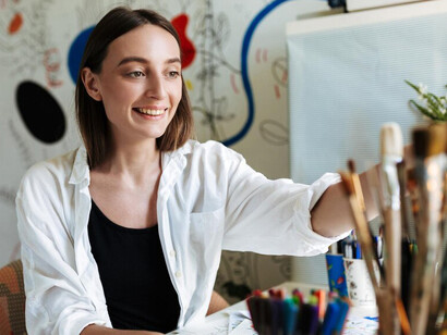 In her home studio, a girl holds paintbrushes while looking thoughtfully at the camera, with a vibrant patterned canvas in the background, reflecting the creative process in her workspace