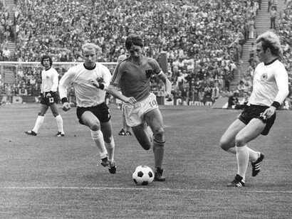 Johan Cruijff (al centro) in azione nella finale del torneo tra i Paesi Bassi e la Germania Ovest (1974). Cruijff è ritratto prima di entrare in area ed essere atterrato al primo minuto di gioco. Si notano Müller (sullo sfondo), Vogts (a sinistra) e Hoeneß che commetterà il fallo da rigore (a destra)