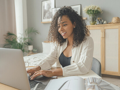 A woman in a white blazer sits at a brown wooden table, working on a MacBook Pro