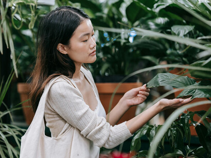 A woman in a white long-sleeve shirt stands among lush green plants, smiling and embracing a simple, happy life