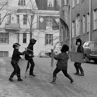 Eeva Rista, Four boys fencing with sticks and shields at the end of Meritullinkatu. Meritullinkatu 32 to the left and Meritullinkatu 33 to the right. Kristianinkatu 7 in the centre, January 1971. Courtesy of Helsinki City Museum