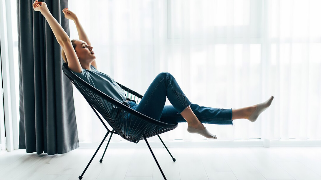 A young woman relaxes in her modern living room, sitting on a stylish chair by the window, embodying the essence of minimalist living