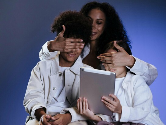 Uma mãe cobre os olhos dos filhos enquanto eles seguram um tablet diante da tela. Em tempos de hiperexposição digital, proteger também significa compreender o que os adolescentes consomem online. O gesto de proteção contrasta com a presença constante das telas no cotidiano infantil. A supervisão digital tornou-se parte essencial do cuidado emocional e cognitivo. Entre curiosidade e vulnerabilidade, crianças e adolescentes acessam conteúdos para os quais muitas vezes ainda não possuem maturidade emocional. O cérebro em formação responde intensamente aos estímulos digitais