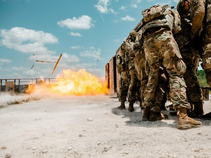 Um grupo de soldados enfrenta uma explosão durante um exercício militar ao ar livre, cercado por poeira e equipamentos de proteção. Entre treinamento, estratégia e tecnologia, a cena remete à permanente preparação das sociedades para conflitos que podem assumir dimensões cada vez mais destrutiva