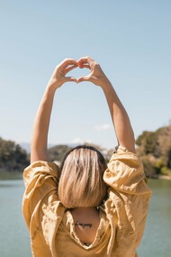 A woman  by a serene lake, her arms extended above her head, forming a heart shape with her hands against a clear blue sky, an image that embodies the balance between nature, self-care, and resilience