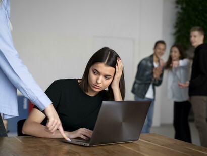 Un giovane donna, visibilmente turbata e con la testa appoggiata alla mano, è seduta davanti al laptop, mentre sullo sfondo altre persone la prendono in giro e ridono