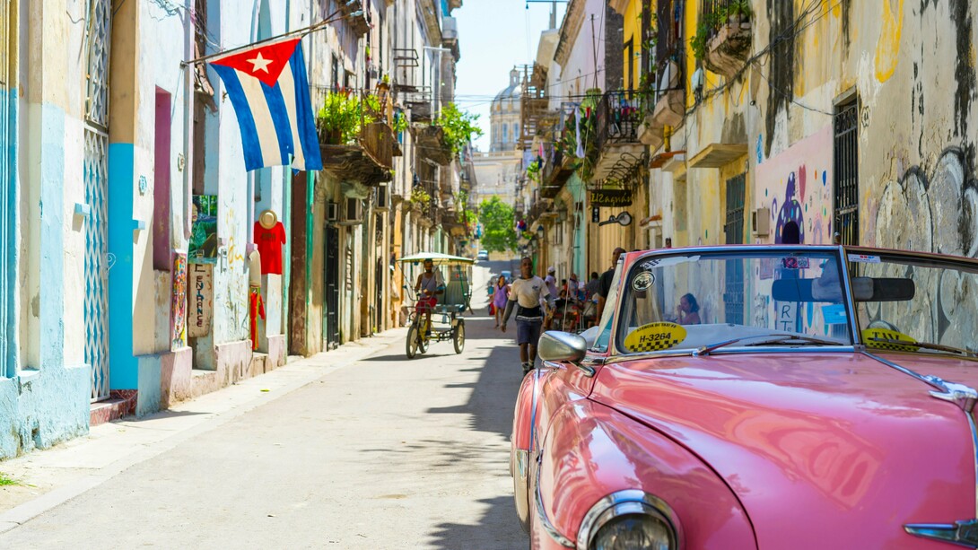 A vintage pink convertible taxi rolls through a colorful Havana street under the Cuban flag, evoking a vivid portrait of resilience, history, and the everyday life shaped by decades of geopolitical tension and economic isolation