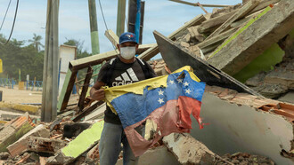 Ciudadano venezolano sostiene una bandera nacional junto a su restaurante dañado tras un terremoto en La Guaira, Venezuela, como signo de resiliencia. 28 de junio de 2026