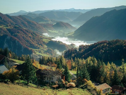 A wide valley unfolds in layered greens and autumn tones, placing the traveler at a distance and emphasizing how small we are within a landscape that has existed long before us