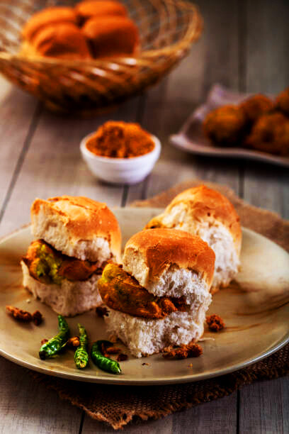 In a restaurant setting, a plate of Mumbai Vada Pav, a deep-fried potato dumpling in a bread bun with green chili and tomato, is presented in the foreground. An unrecognizable person picks up a slice of tomato, with the focus on the vibrant dish