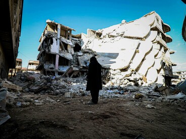A person stands in front of a demolished building in an urban area, Syria