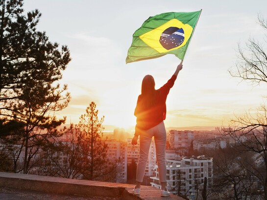Uma mulher de calça branca e camisa vermelha segura a bandeira brasileira diante de uma cidade iluminada pelo sol. Mais do que um símbolo nacional, a cena sugere a presença feminina como parte ativa das escolhas que definem os caminhos do Brasil