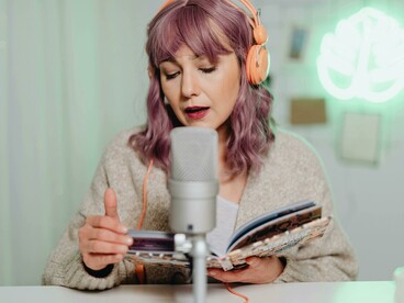 A podcast host reads from a book during a casual indoor recording session, wearing headphones and speaking into a microphone