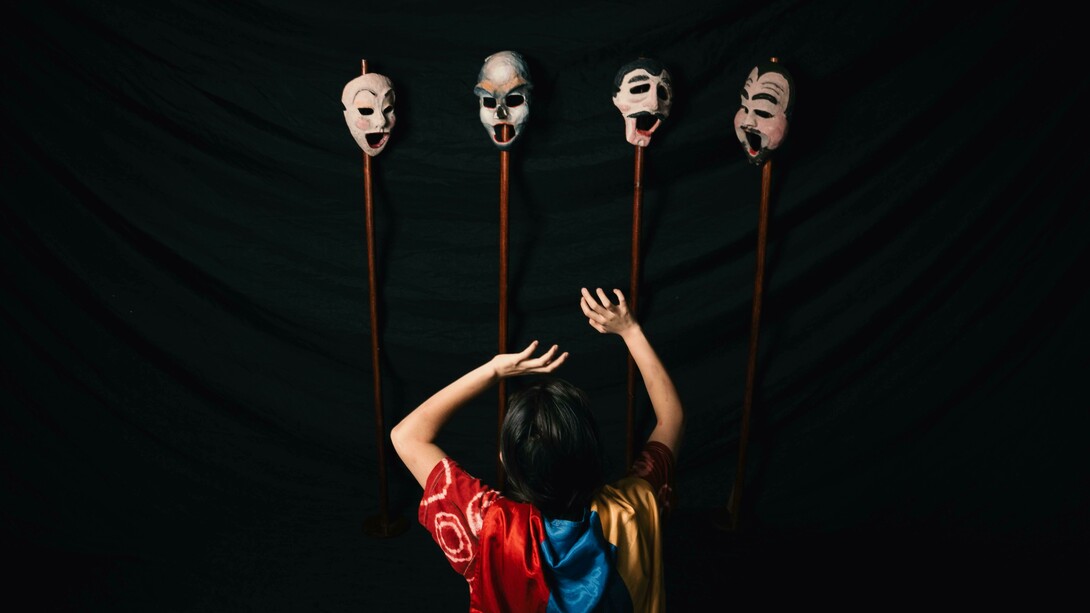 A woman in a robe performs with theater masks against a black background, embodying the tension between identity and performance