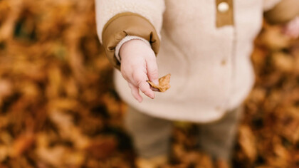 Un bambino con un cappotto autunnale prende tra le mani una foglia arancione tipica del mese di Ottobre